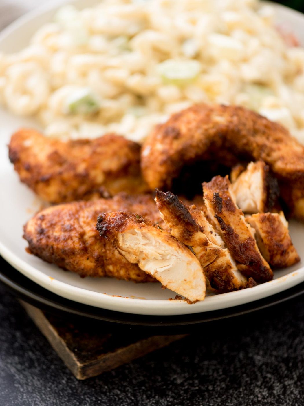 Air fryer chicken tenders rest on a white plate alongside a serving of creamy pasta salad, all elegantly placed on a dark surface.