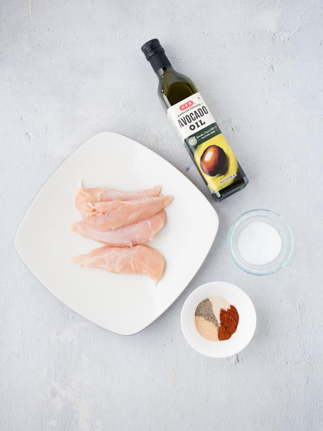 Raw chicken breasts on a square plate, ready to be transformed into satisfying air fryer chicken tenders. Accompanied by a bottle of avocado oil, a small bowl of salt, and a spice blend featuring paprika, pepper, and garlic powder, all beautifully arranged on a light gray surface.