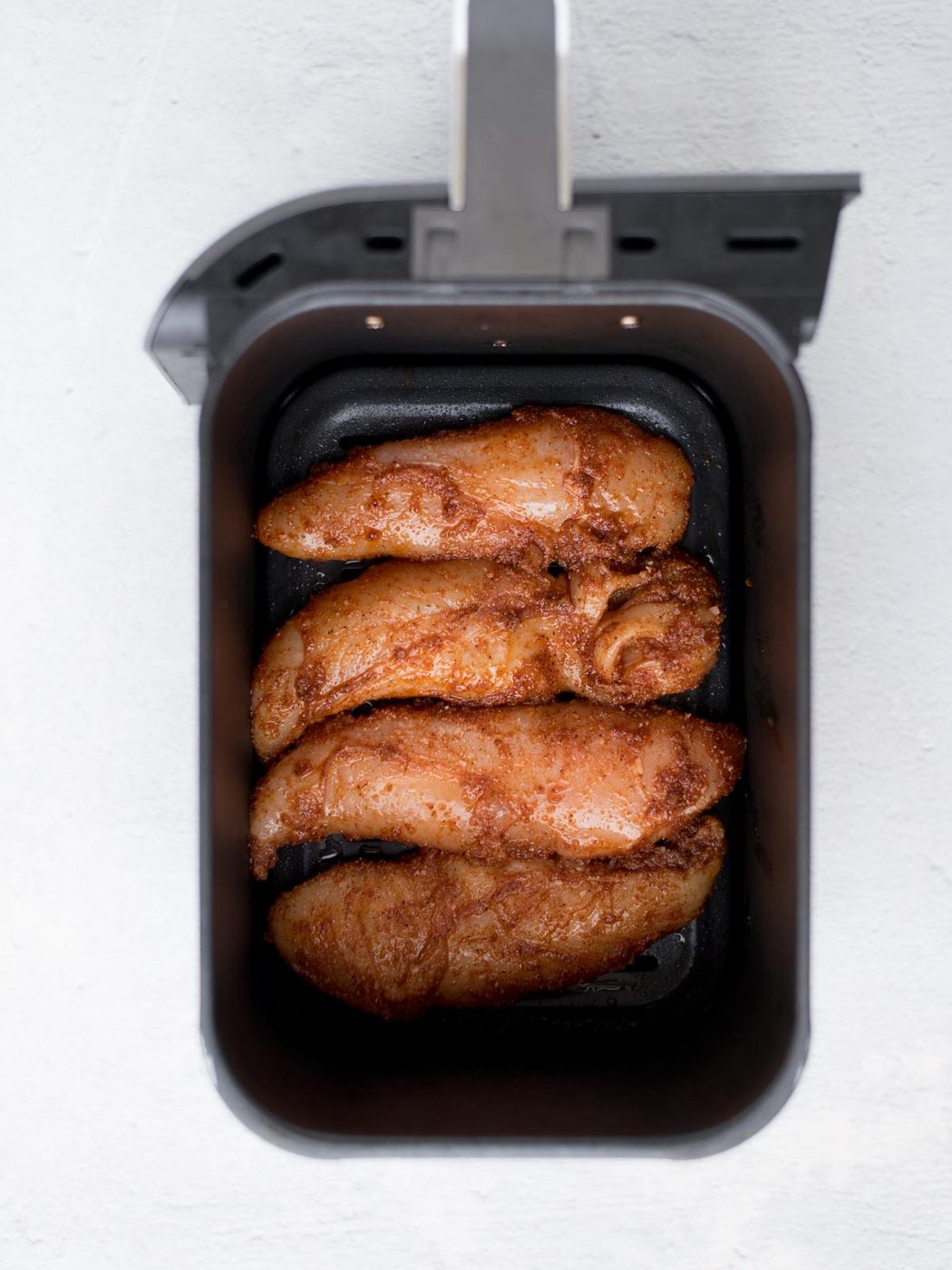 Four seasoned air fryer chicken tenders rest in the basket, viewed from above on a light surface. They appear golden brown, evenly cooked or recently fried, and ready to be enjoyed.