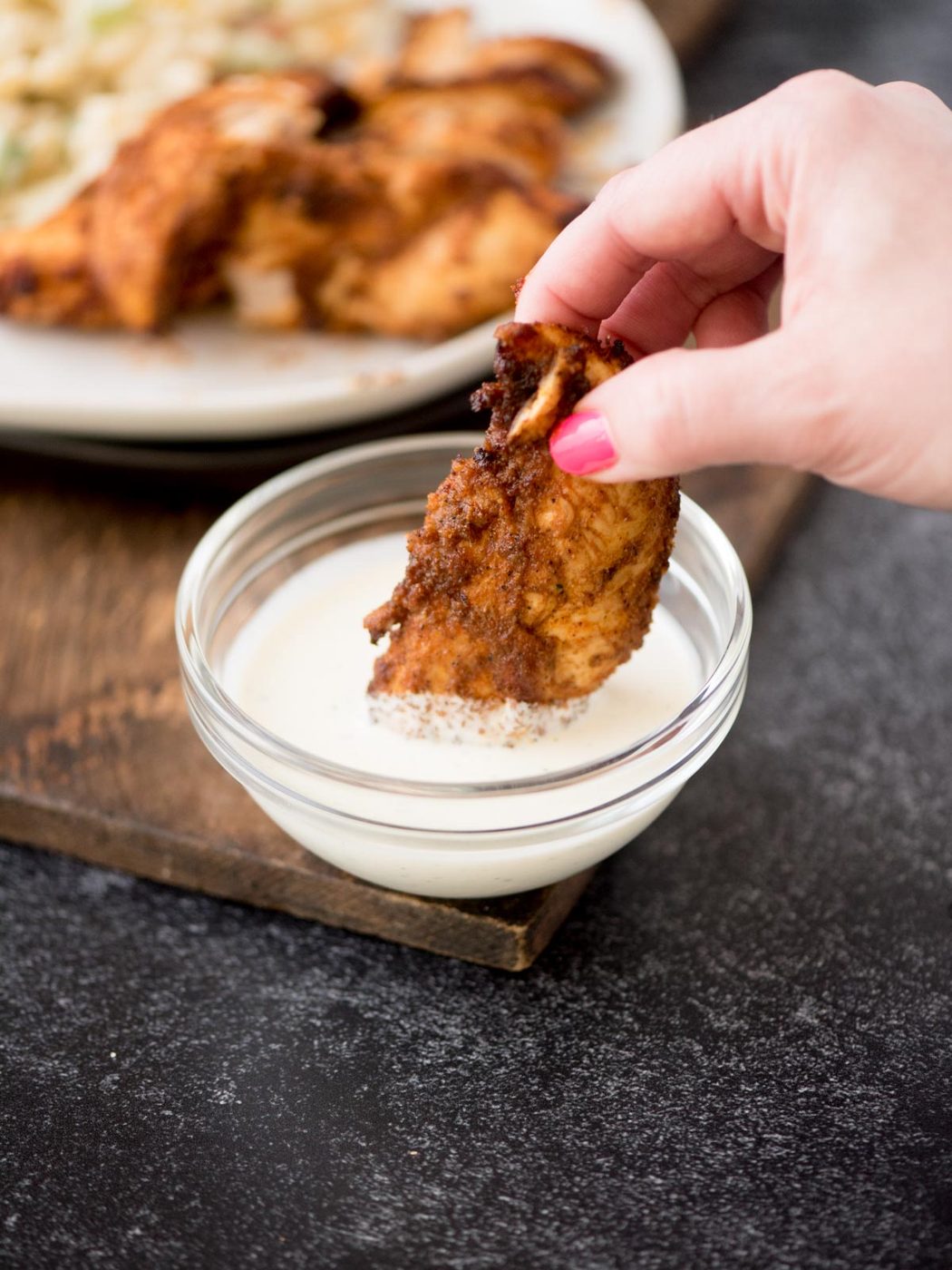 A hand with pink nail polish dips a piece of seasoned, cooked air fryer chicken tender into a small clear bowl of white sauce. In the background, theres a wooden board with more chicken tenders on a white plate. The surface is dark.