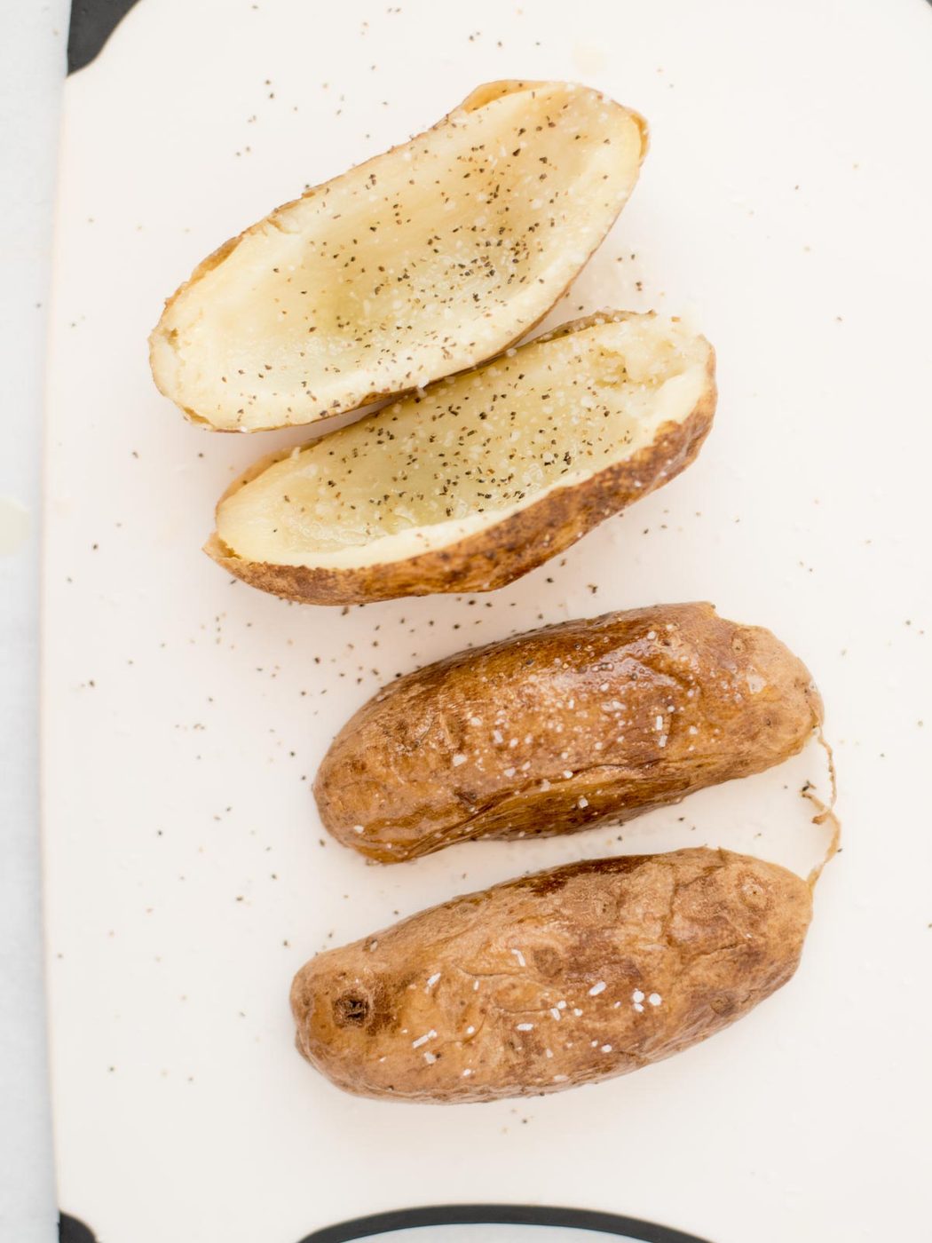 4 russet potatoes, 2 upright, 2 upside down, laying on parchment paper, being sprinkled with salt and pepper on both sides of the potato.