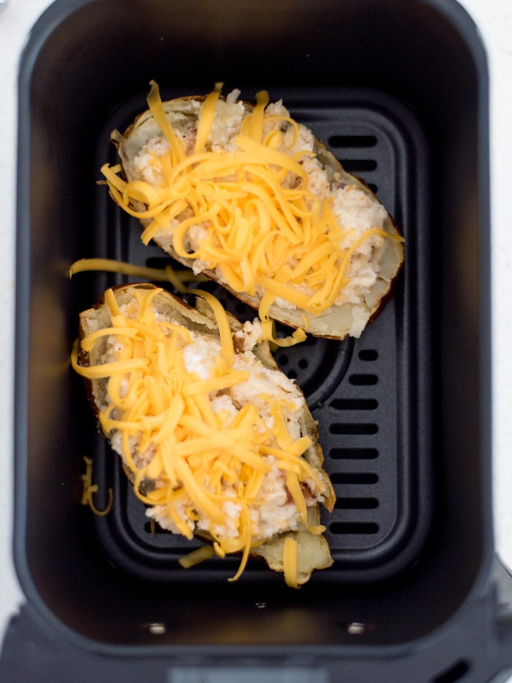 Two twice baked potatoes topped with shredded cheese in the air fryer ready to cook.