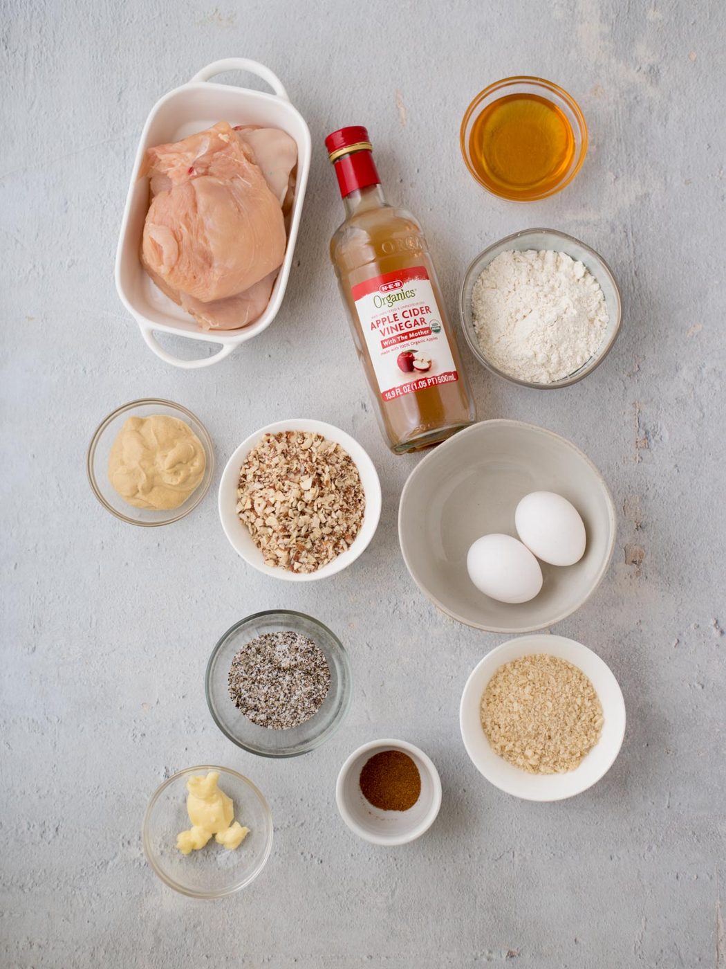 Various ingredients are arranged on a light surface, including raw chicken ready for a crispy almond crusted twist, apple cider vinegar, mustard, eggs, flour, herbs, honey, butter, nuts, seeds, and breadcrumbs in bowls and containers.
