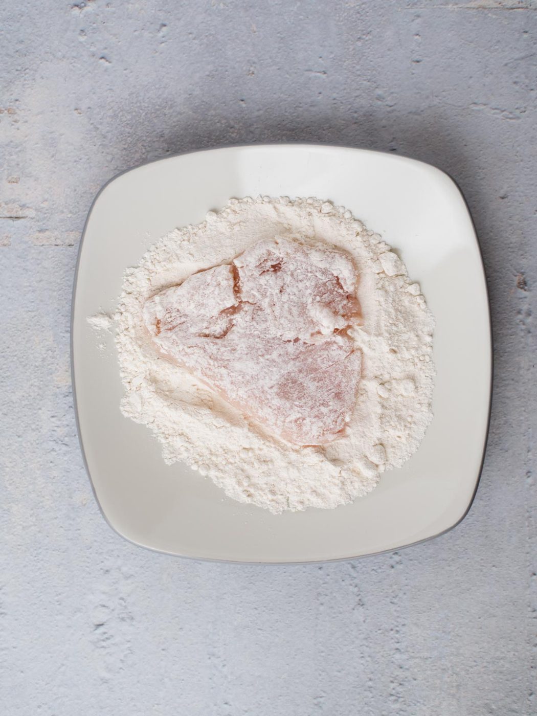 A piece of raw meat, destined to become almond chicken, is coated in flour and rests on a square white plate. The plate sits elegantly on a textured, light gray surface.