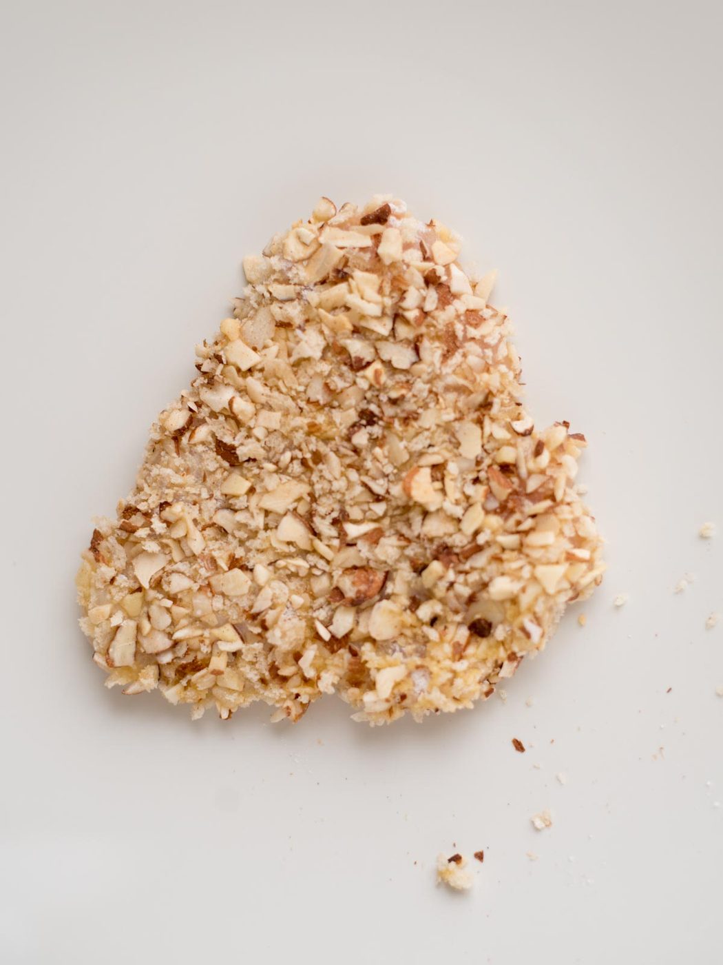 A heart-shaped food item coated with a crunchy almond crust, resting on a plain white surface. Some nut crumbs are scattered around it.