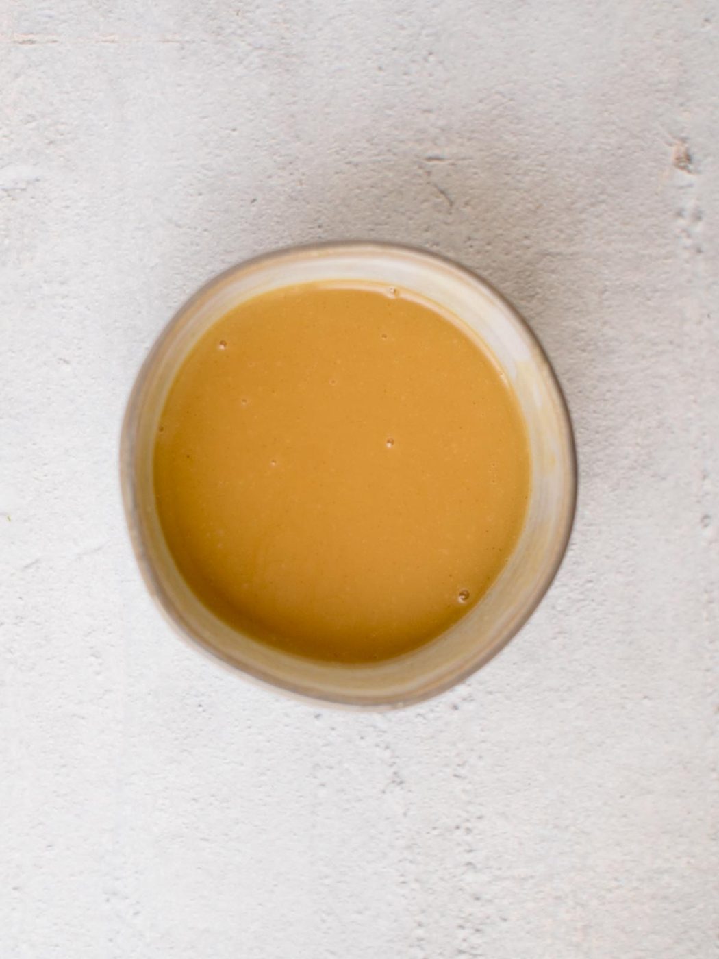 A round bowl filled with smooth, light brown sauce sits on a textured gray surface, evenly complementing the flavors of crispy almond chicken.