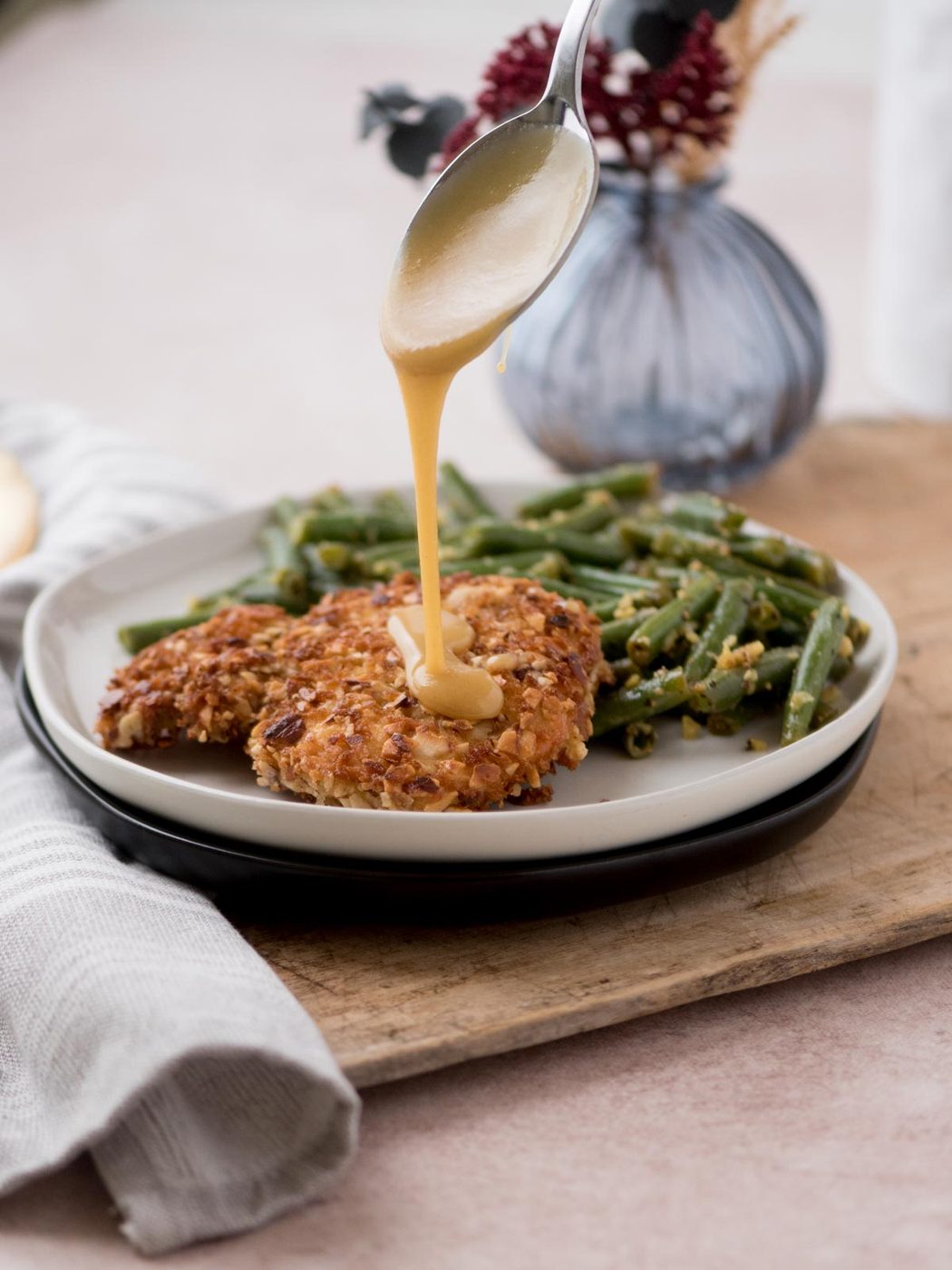 A spoon drizzles gravy over a piece of crispy almond chicken served on a white plate with green beans. A gray napkin and decorative vase with flowers are in the background.