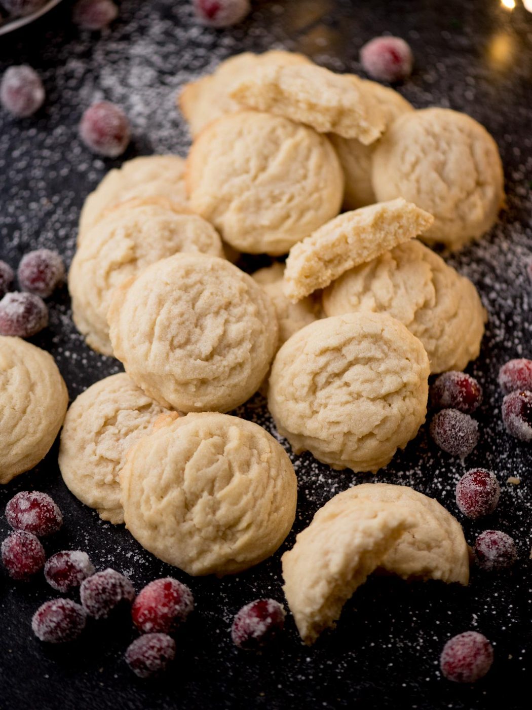 A pile of shortbread cookies is scattered on a dark surface, surrounded by sugared cranberries and a dusting of powdered sugar. Some cookies are whole, while a few are broken, revealing their crumbly texture reminiscent of a house favorite Amish sugar cookies.