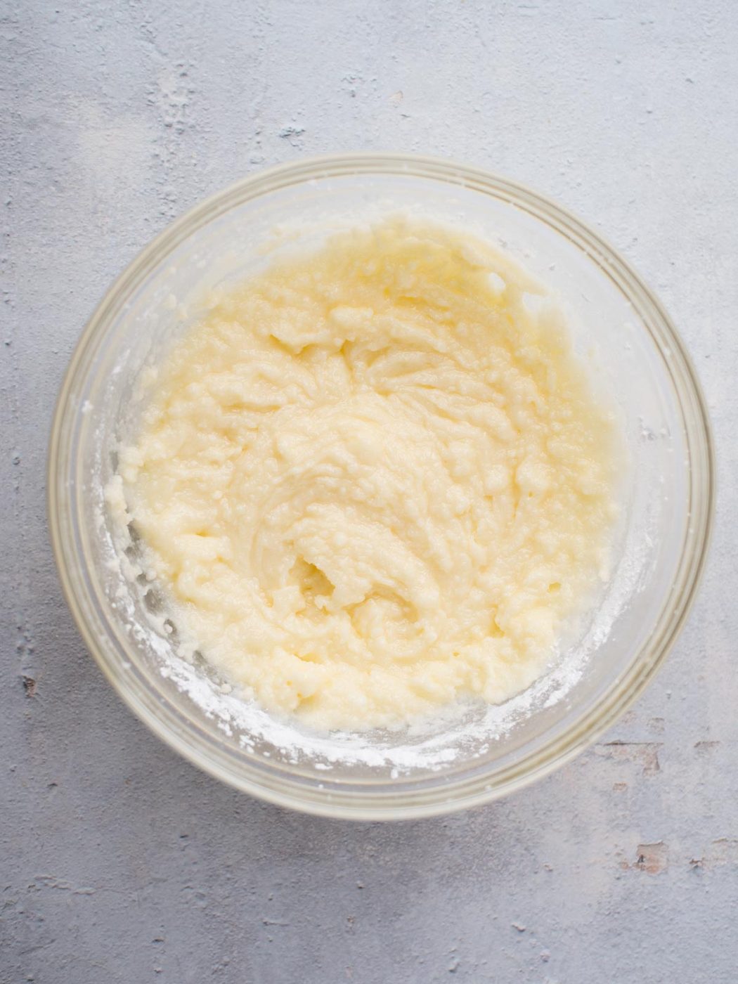 A clear glass bowl filled with a creamy, light yellow batter sits on a textured gray surface. The smooth mixture, with its few small lumps, promises the delectable taste of classic Amish sugar cookies in every bite.