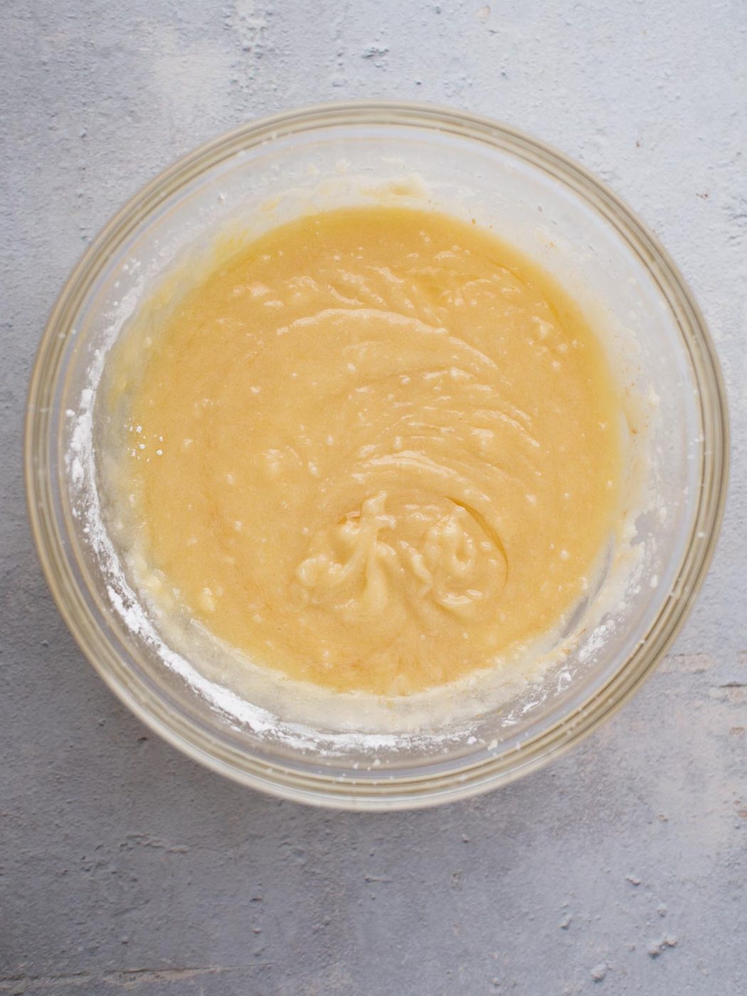 A glass bowl containing smooth, light yellow cake batter sits on a textured gray surface. The batter is slightly swirled in the center, resembling the rich mixture of Amish sugar cookies that have just been expertly combined.