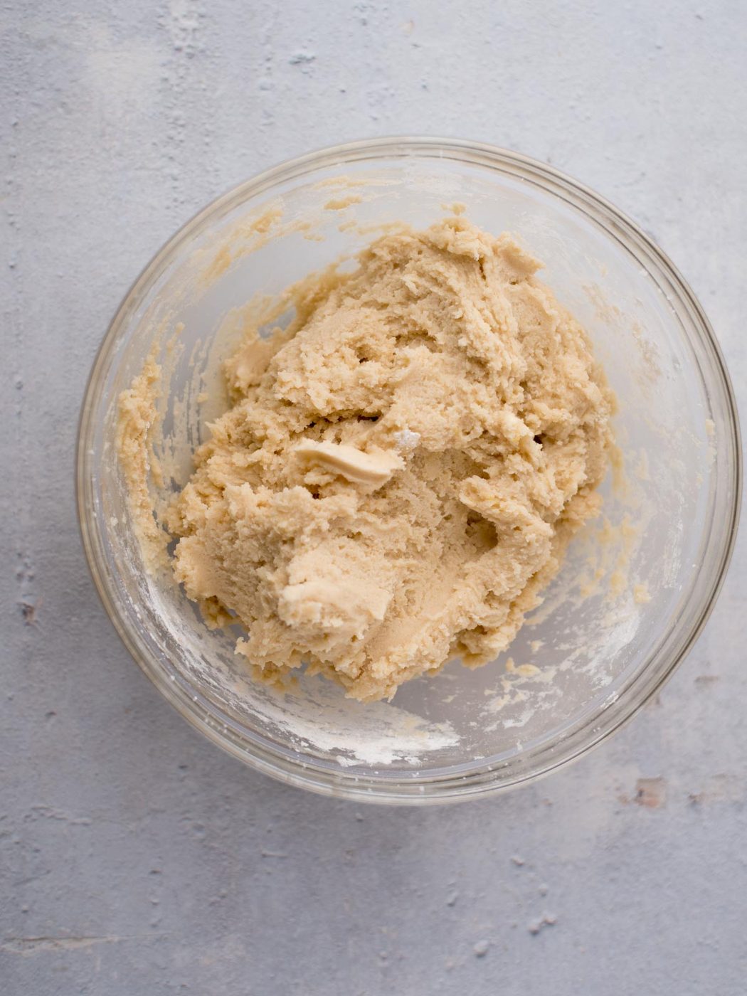 A glass bowl filled with unbaked Amish sugar cookie dough sits on a textured light gray surface. The dough appears creamy and thick, mixed to a smooth consistency that promises a house favorite sugar cookies.