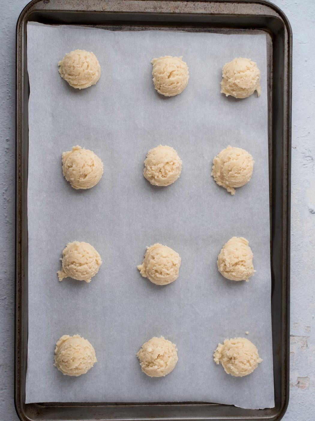Twelve dough balls, suited to a house favorite Amish sugar cookies, are evenly spaced on a parchment-lined baking sheet, ready to be baked.