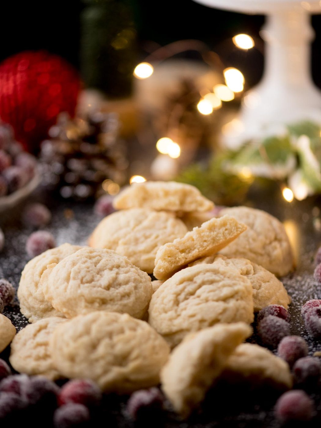 A pile of a house favorite sugar cookies is surrounded by sugared cranberries on a festive table. The background features blurred twinkling lights and a red ornament, creating a cozy holiday atmosphere.