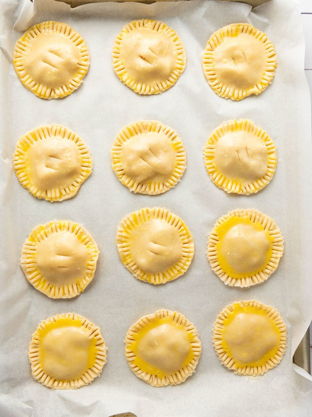 Twelve round unbaked hand pies arranged in a grid on a parchment-lined baking sheet.
