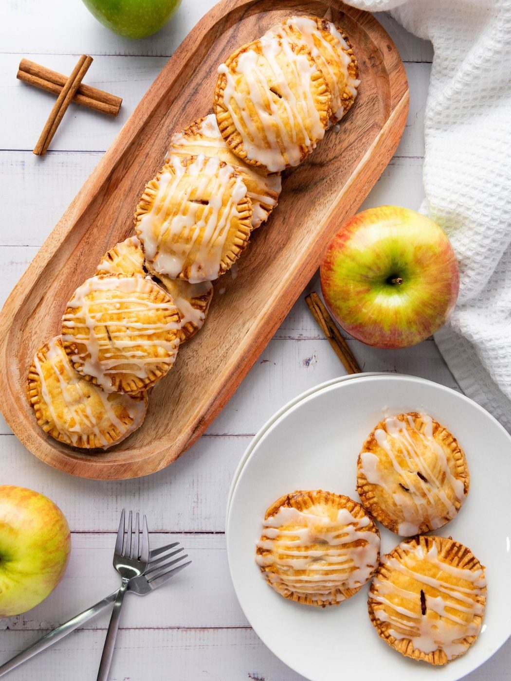 Mini apple pies with white icing arranged on a wooden platter and a white plate, accompanied by fresh apples and cinnamon sticks.