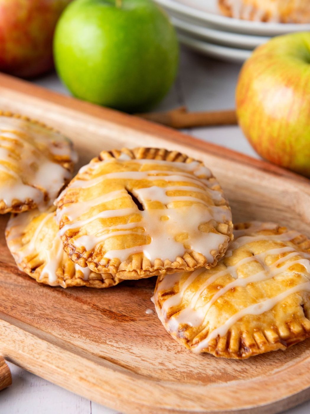 Three small, round hand pies drizzled with white glaze on a wooden board, with two whole apples in the background.