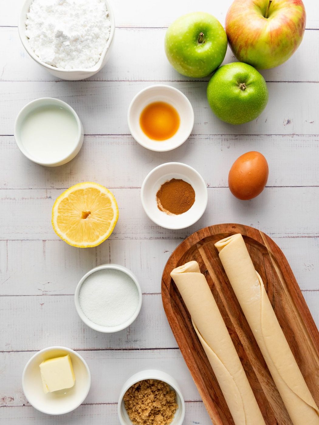 Ingredients for a pastry recipe, including apples, dough, an egg, lemon, sugar, cinnamon, butter, milk, and vanilla extract, arranged on a wooden surface.