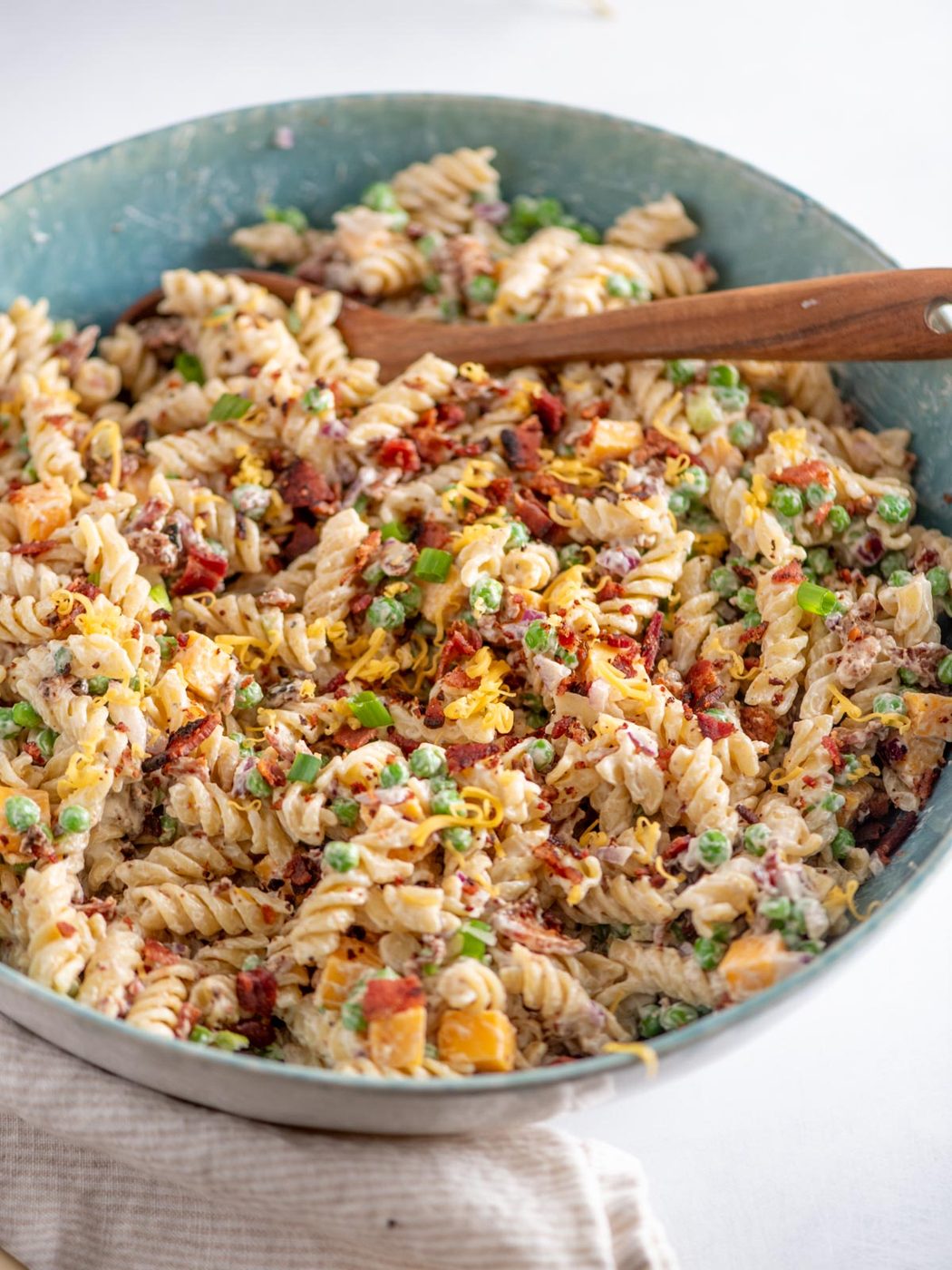 A large bowl of creamy rotini pasta mixed with peas, sun-dried tomatoes, chopped vegetables, and herbs, topped with shredded cheese, with a wooden serving spoon resting in the bowl.
