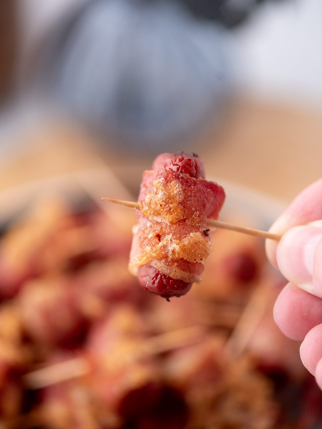 A hand holds a toothpick skewering a small sausage wrapped in crispy bacon, with more bacon-wrapped sausages blurred in the background.