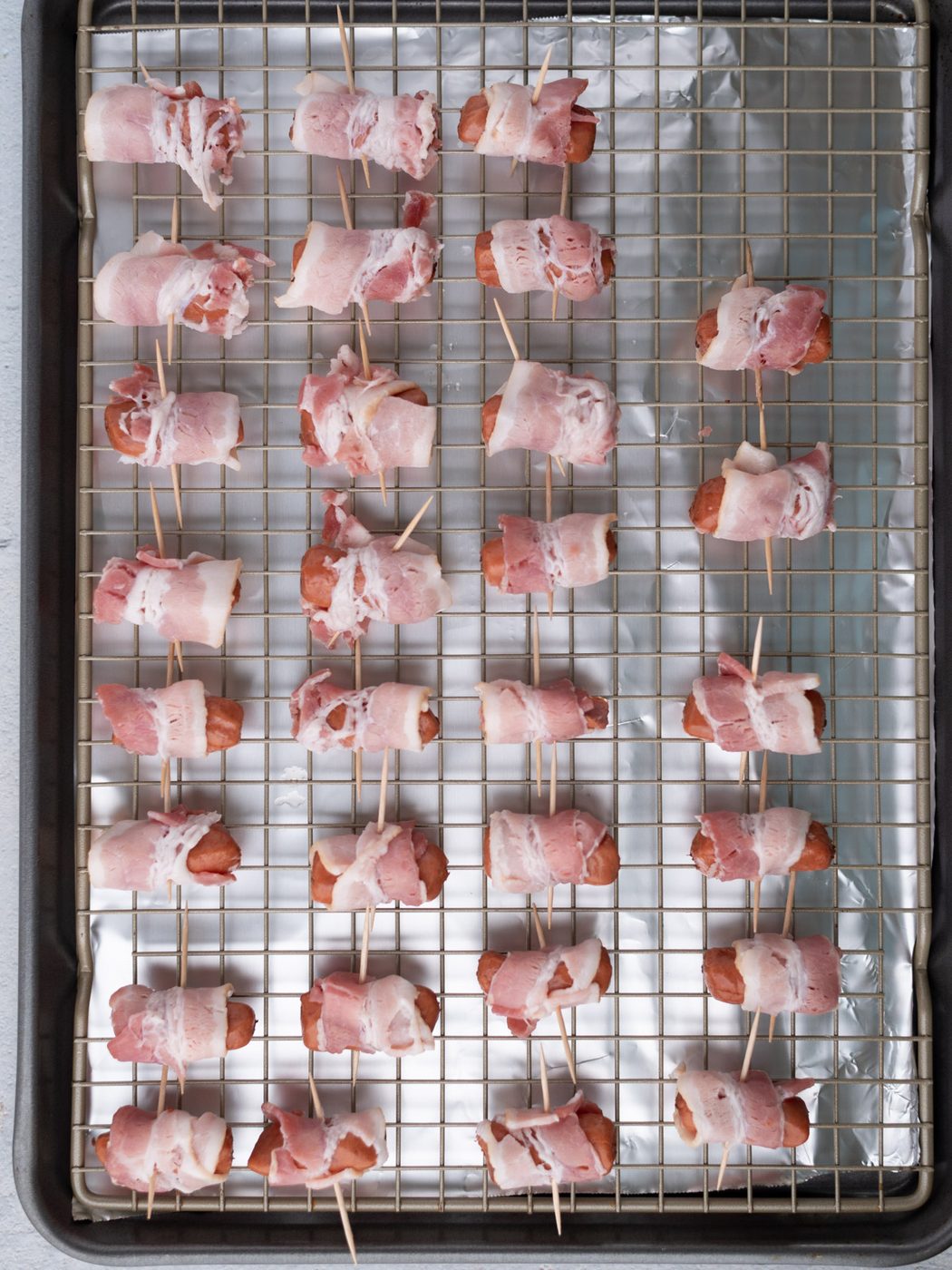 Raw bacon-wrapped little smokies skewered with toothpicks are arranged in rows on a wire rack over a foil-lined baking sheet, ready to be cooked.
