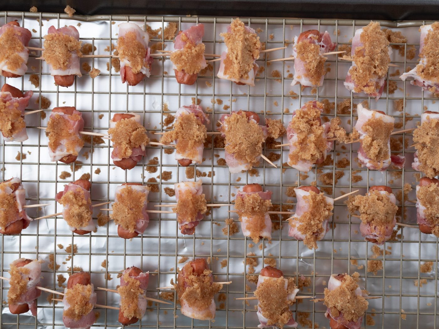 Raw bacon-wrapped bites on a wire rack, topped with brown sugar, arranged in rows and secured with toothpicks, ready for baking on a foil-lined baking sheet.