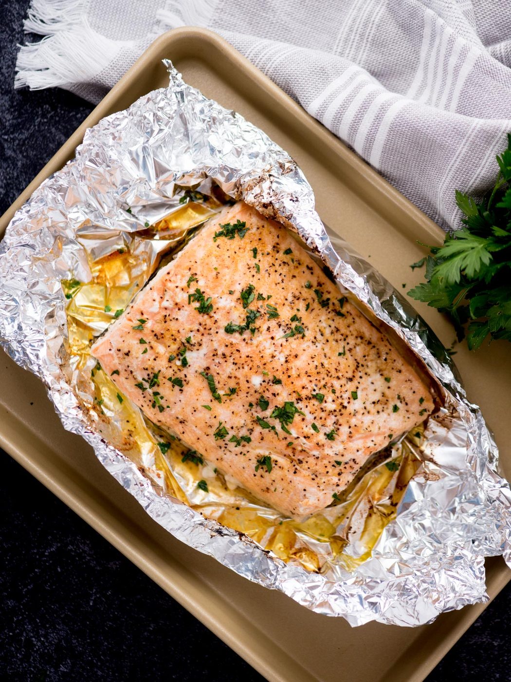 baked salmon in foil on a baking sheet surrounded by fresh parsley and a kitchen towel