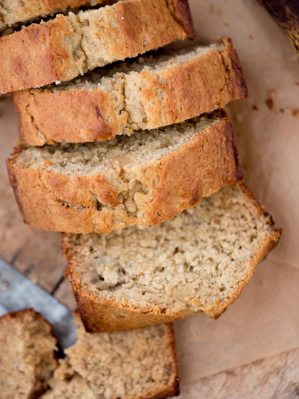 Sliced banana bread on a wooden surface, showing a moist and dense texture. The golden-brown crust suggests its freshly baked. A knife is partially visible at the edge.