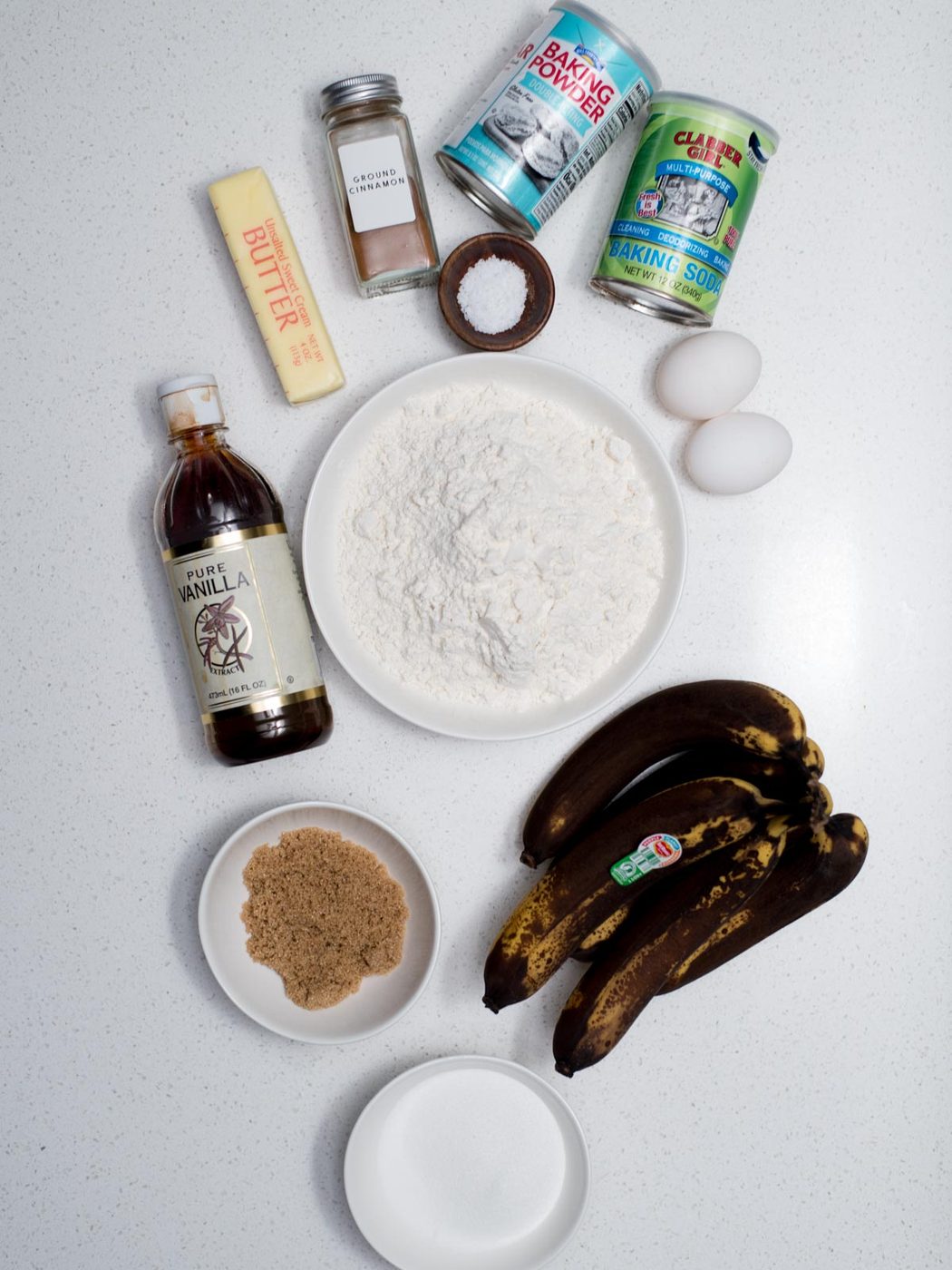 Ingredients for baking on a white surface: brown bananas, flour in a bowl, eggs, a stick of butter, vanilla extract, brown sugar, baking powder, baking soda, cinnamon, and salt.