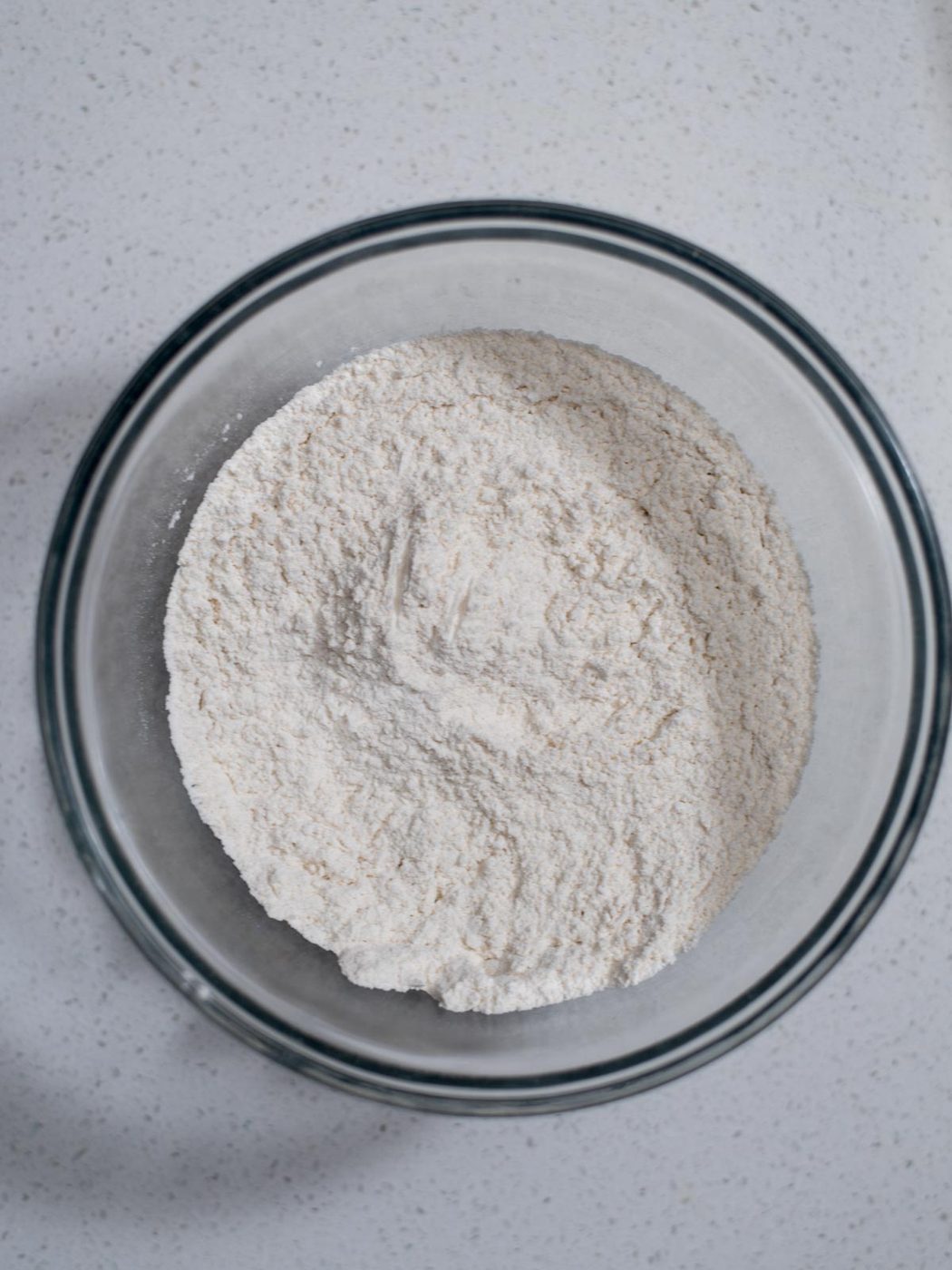 A glass bowl filled with white flour sits on a speckled white countertop. The flour appears finely milled and is slightly mounded in the center of the bowl.