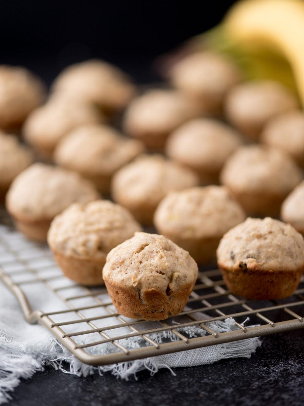 Mini banana muffins are cooling on a wire rack, with several muffins in sharp focus at the front and more blurred in the background. The scene suggests a fresh batch just baked and ready to eat.