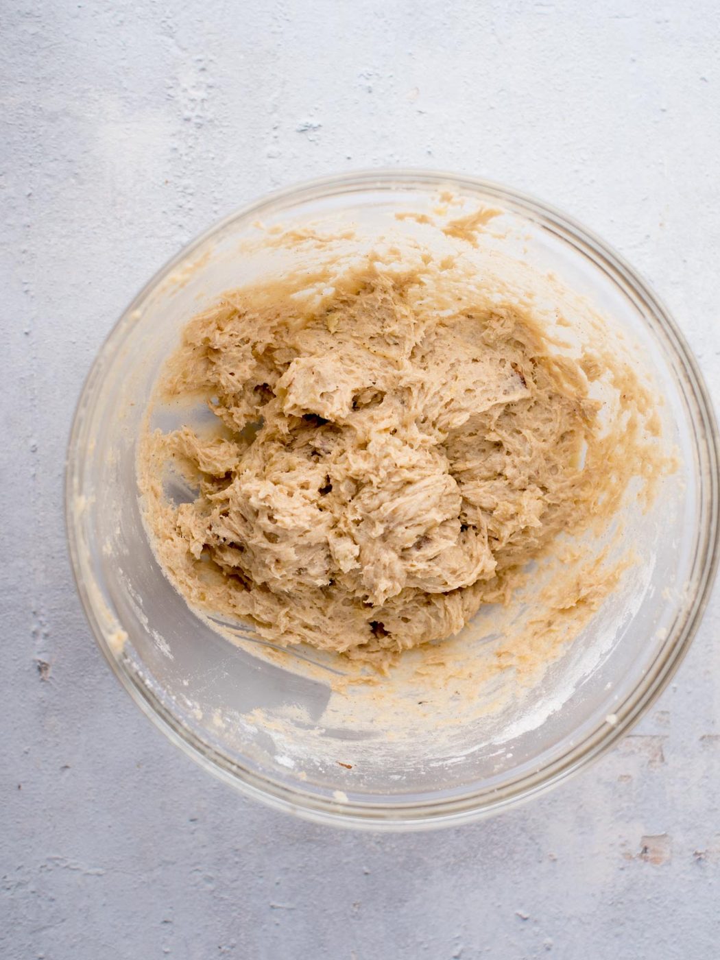 A glass bowl containing thick, creamy cookie dough sits on a light-colored, textured surface. The dough appears partially mixed, with a slightly lumpy texture.