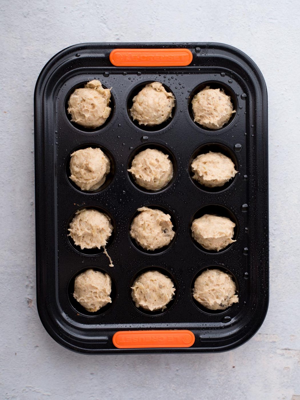 A black muffin tin with twelve round dough balls placed in each cup, ready for baking, sits on a light gray surface. The tin has orange silicone grips on the short ends.