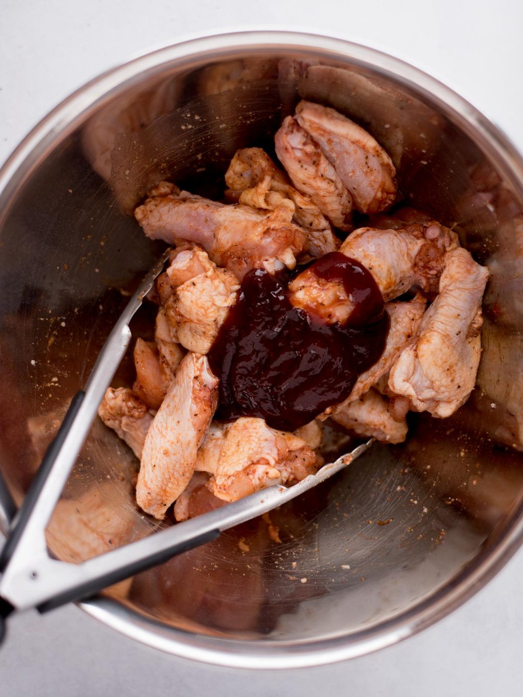 bbq sauce on top of seasoned chicken wings in a stainless steel mixing bowl with tongs