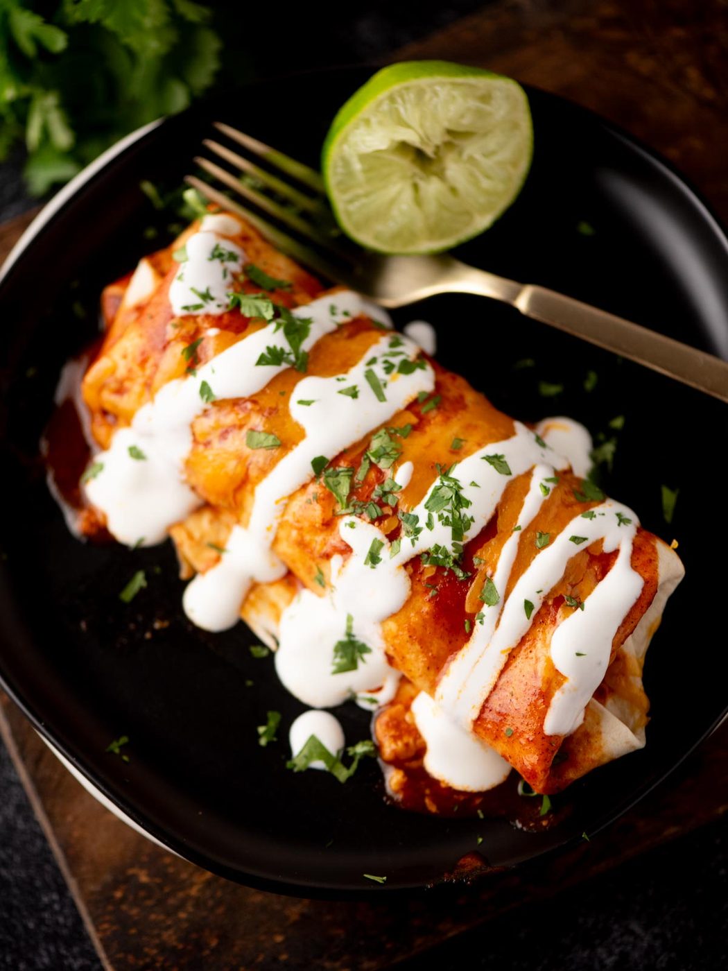 A plate with enchiladas topped with white sauce and chopped herbs, served with a gold fork and a halved lime on the side.