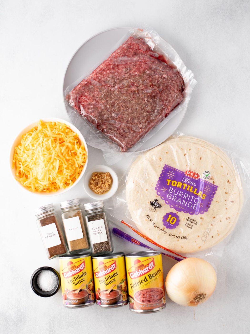 A flat lay of ingredients for burritos: ground beef on a plate, shredded cheese, three cans of refried beans, flour tortillas, an onion, a packet of taco seasoning, minced garlic, and four spice jars.