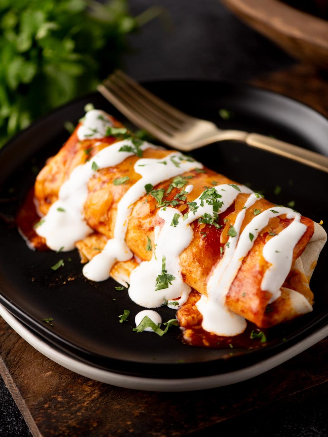 A burrito covered in red sauce and drizzled with white crema, garnished with chopped cilantro, sits on a black plate with a gold fork.