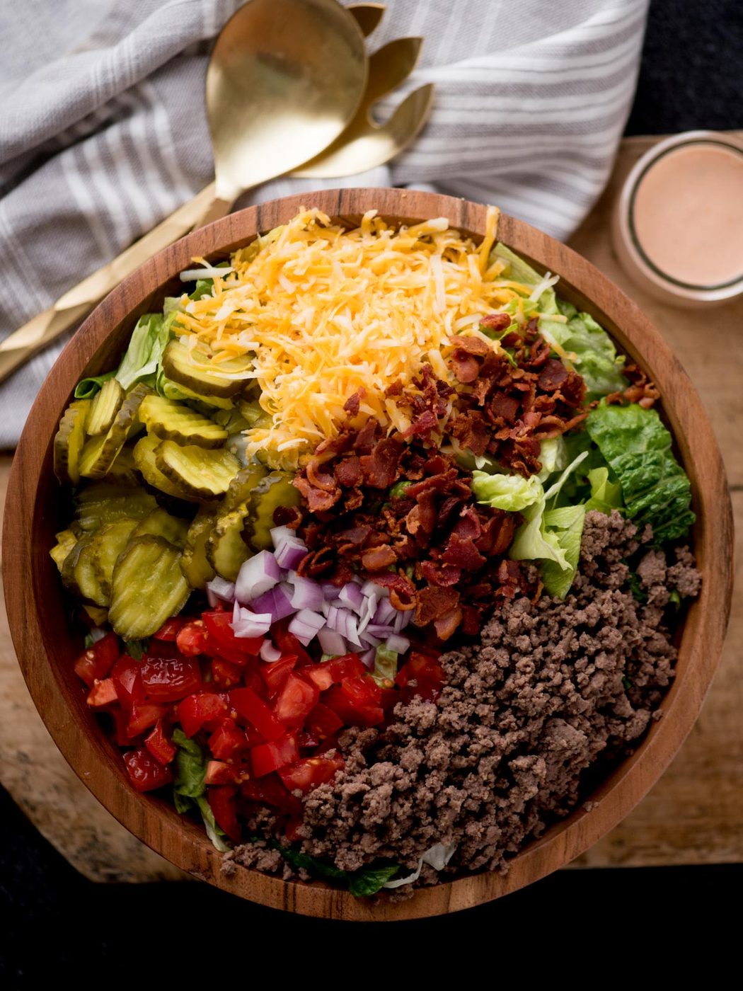 big mac salad in a wooden bowl sitting on a dough board. There are serving utensils, a jar of dressing, and a kitchen towel surrounding it.