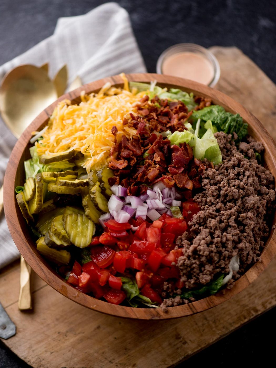 big mac salad in a wooden bowl sitting on a dough board. There are serving utensils, a jar of dressing, and a kitchen towel surrounding it.