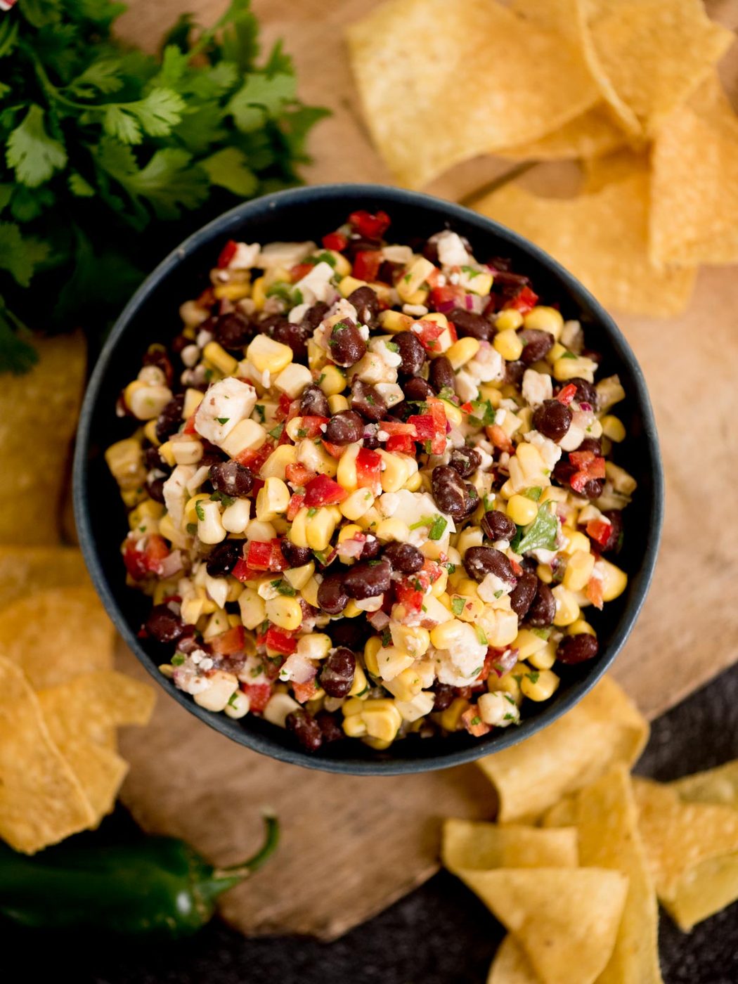 A bowl of colorful black bean corn feta dip with diced red peppers, surrounded by tortilla chips on a wooden surface.