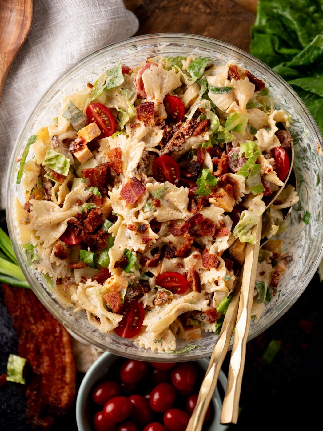 A glass bowl filled with bowtie pasta salad mixed with bacon, lettuce, cherry tomatoes, and a creamy dressing. Two gold utensils rest in the bowl, and fresh greens and tomatoes are nearby.