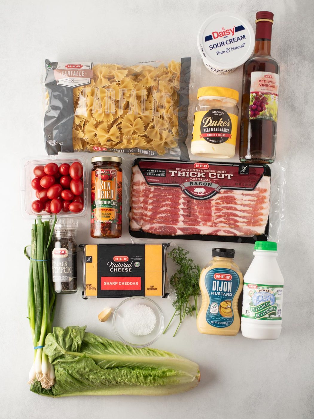A flat lay of ingredients including bowtie pasta, sour cream, red wine vinegar, grape tomatoes, sun-dried tomatoes, thick cut bacon, sharp cheddar cheese, Dijon mustard, whole milk, green onions, fresh dill, Romaine lettuce, garlic, black pepper, and flour.