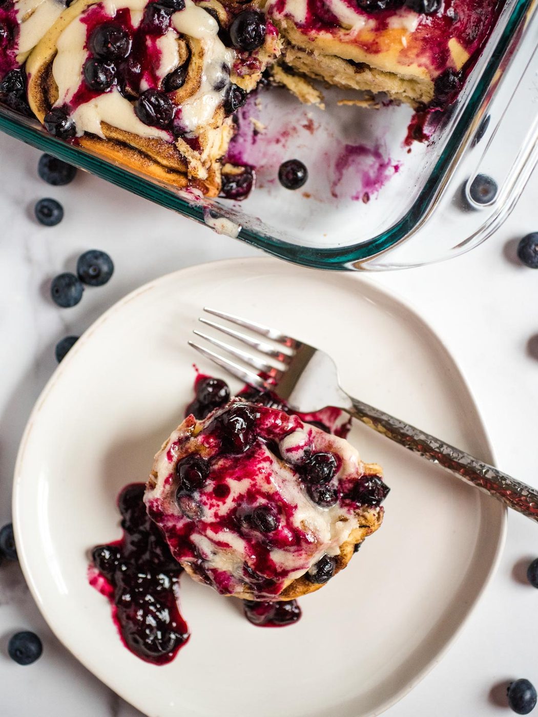 overhead shot of blueberry cinnamon rolls with one scooped out onto a white plate with a fork