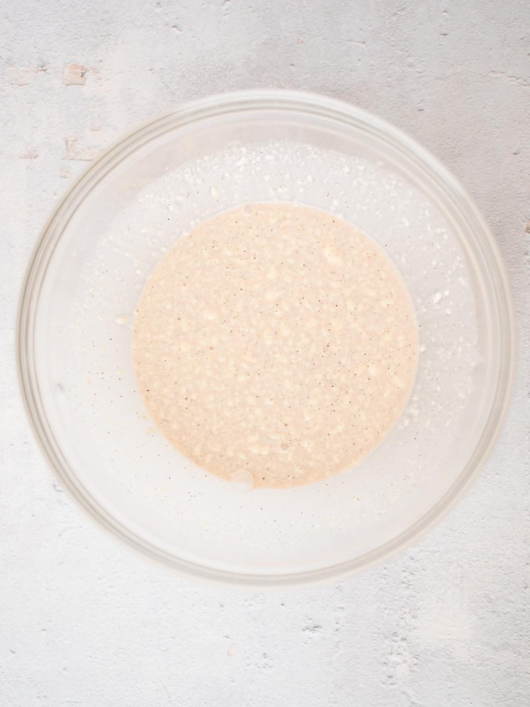 A clear glass bowl containing a bubbly, creamy beige sourdough starter on a light-colored surface. The texture is slightly thick with small air bubbles visible throughout.