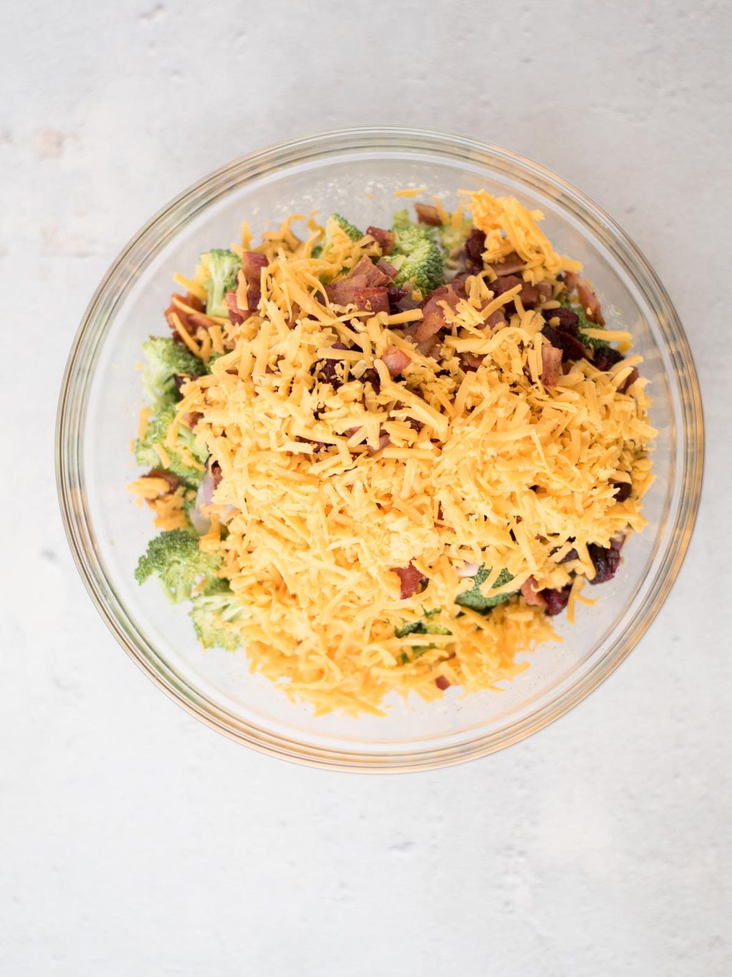 A clear glass bowl filled with a salad of chopped broccoli, crispy bacon bits, raisins, and shredded cheddar cheese, set against a light background.