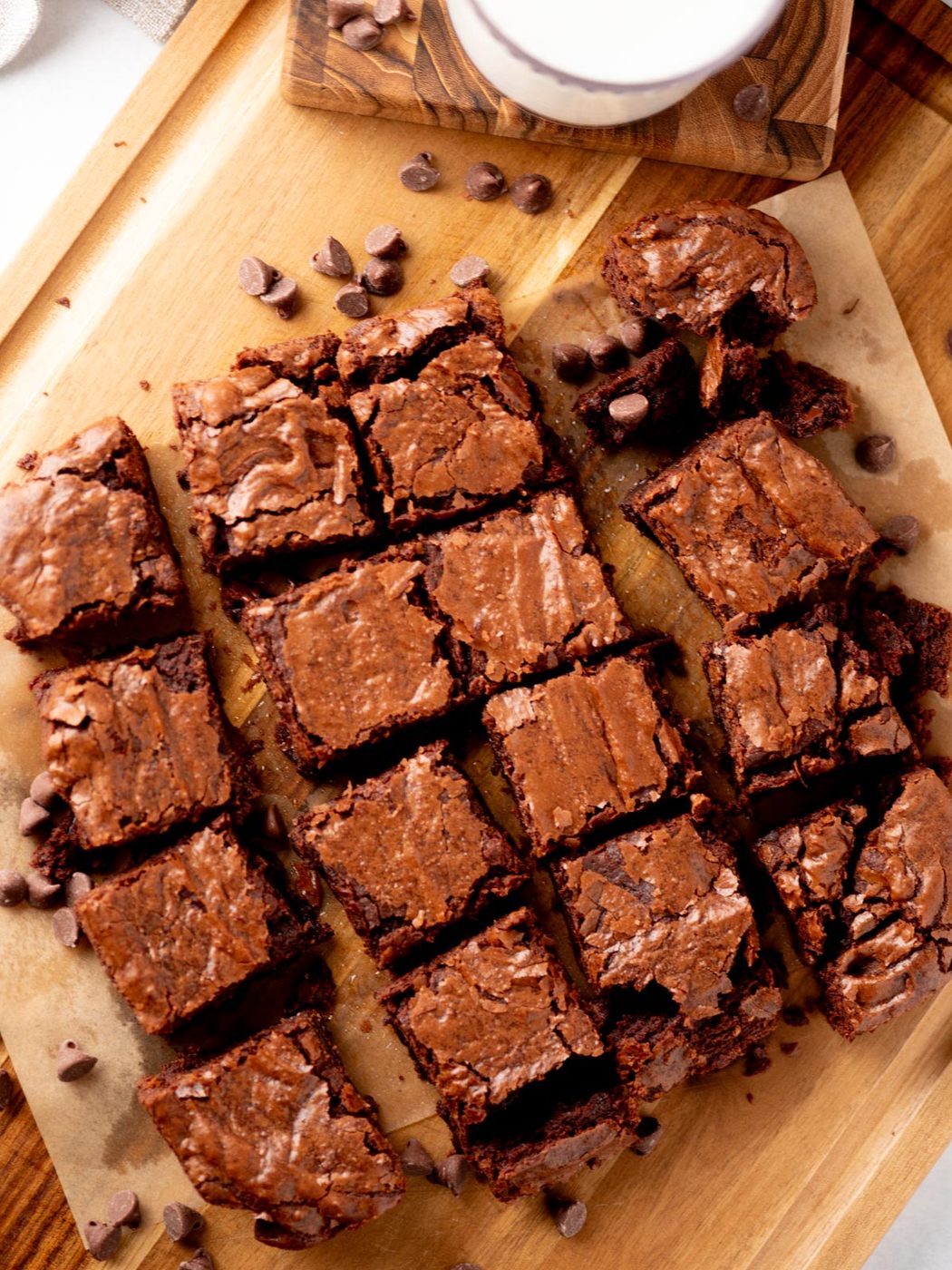 Freshly baked chocolate brownies cut into squares are arranged on parchment paper atop a wooden board, surrounded by scattered chocolate chips. A glass of milk sits nearby.