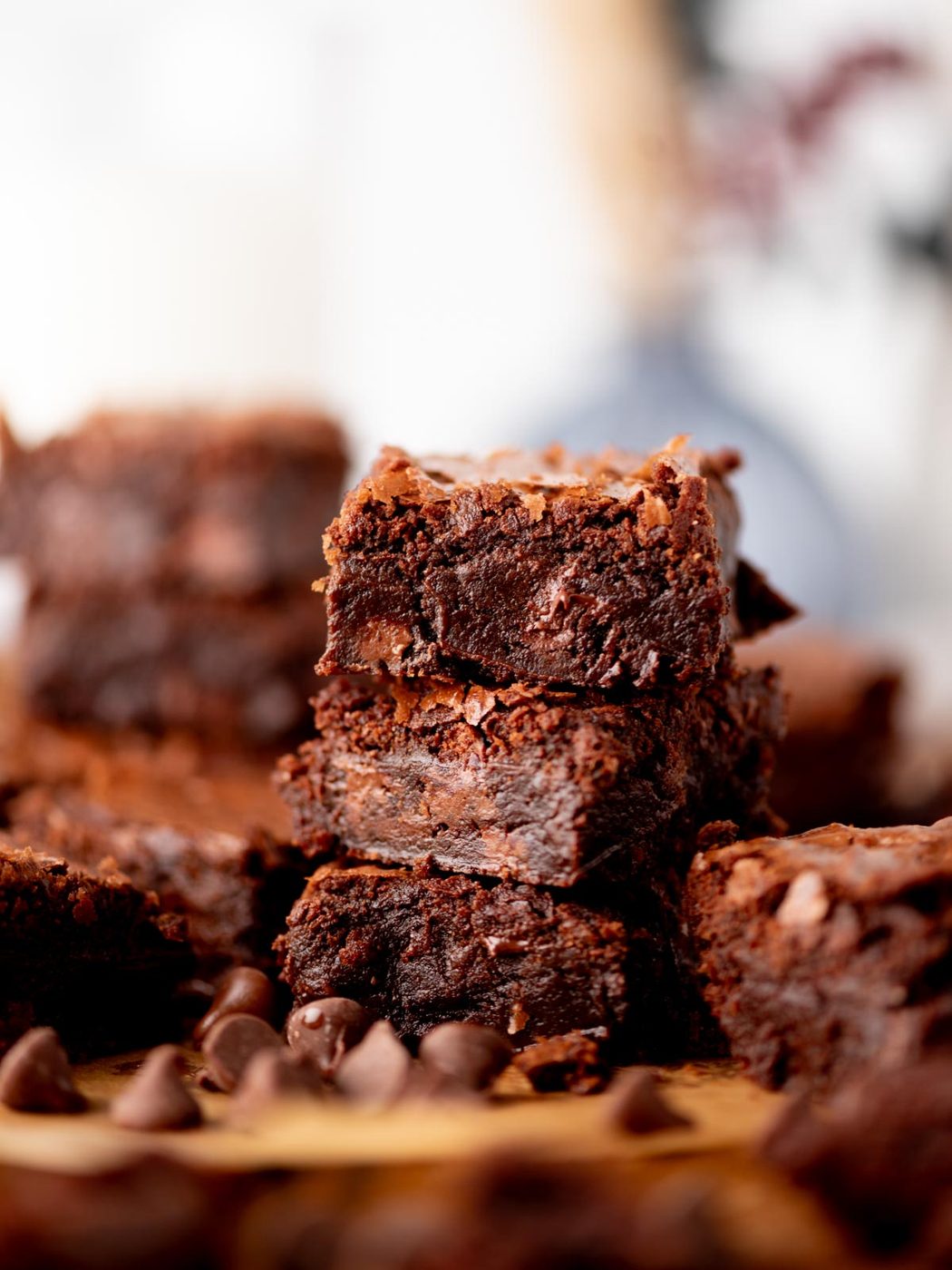 A close-up of a stack of rich, fudgy chocolate brownies with a bite taken out of the top brownie, surrounded by chocolate chips, with more brownies blurred in the background.