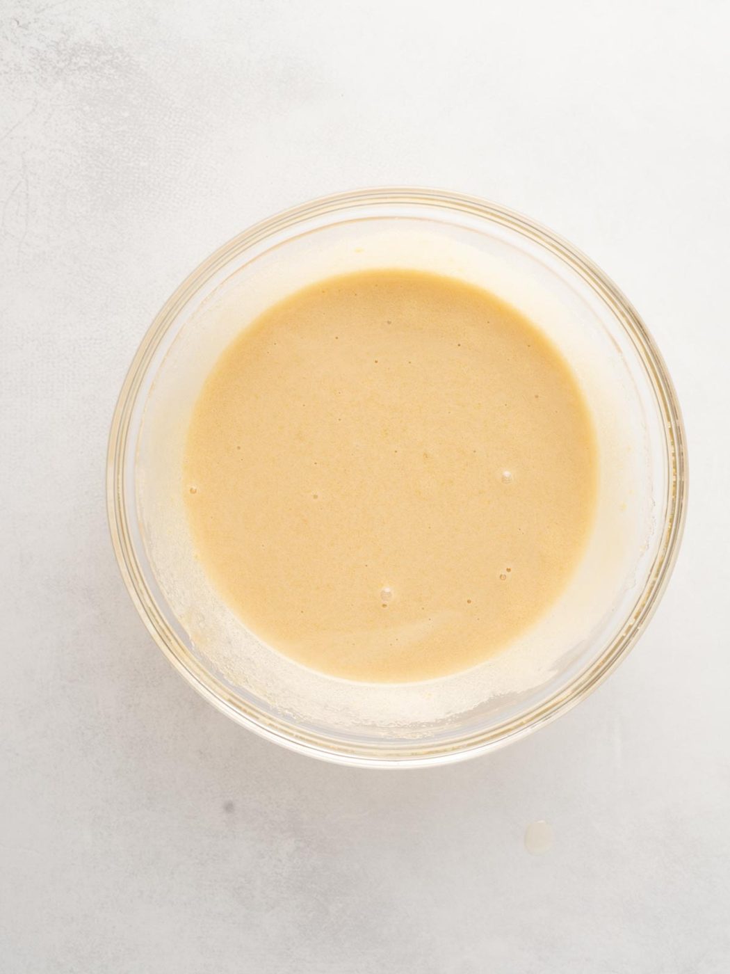 A clear glass bowl filled with a smooth, light tan batter sits on a white surface.