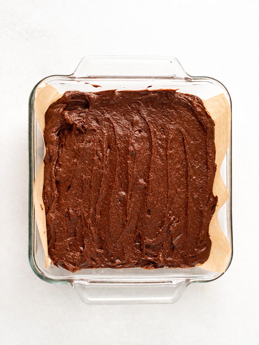 A glass baking dish lined with parchment paper is filled with unbaked chocolate brownie batter, spread evenly across the surface.