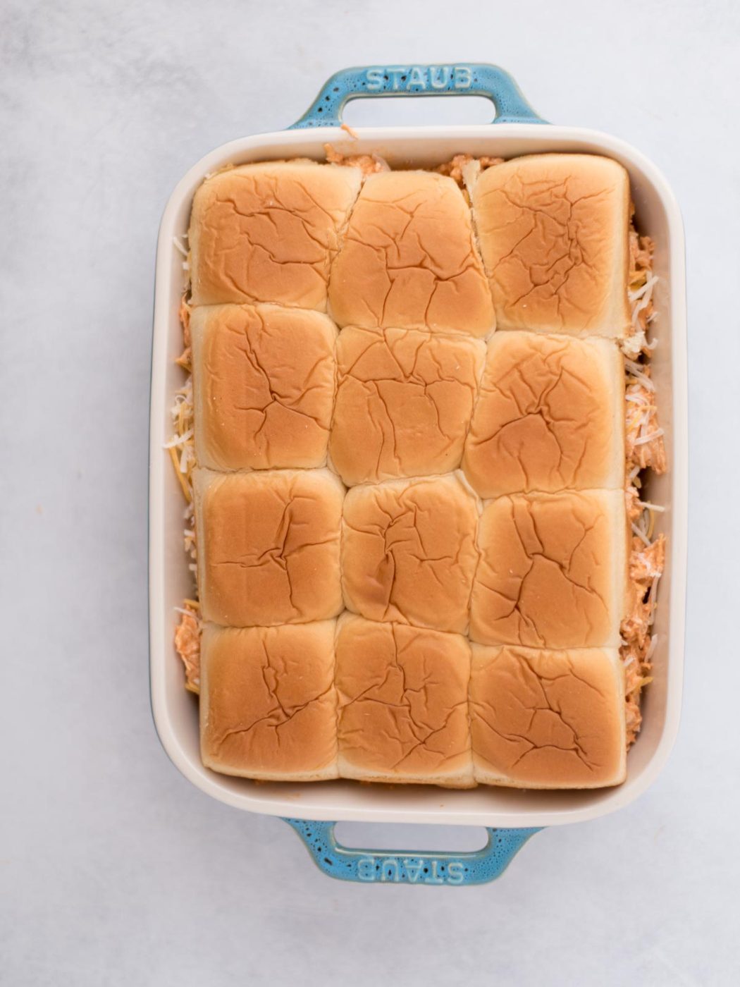 Sliders rolls topped with seasoned meat and shredded cheese in a baking dish.