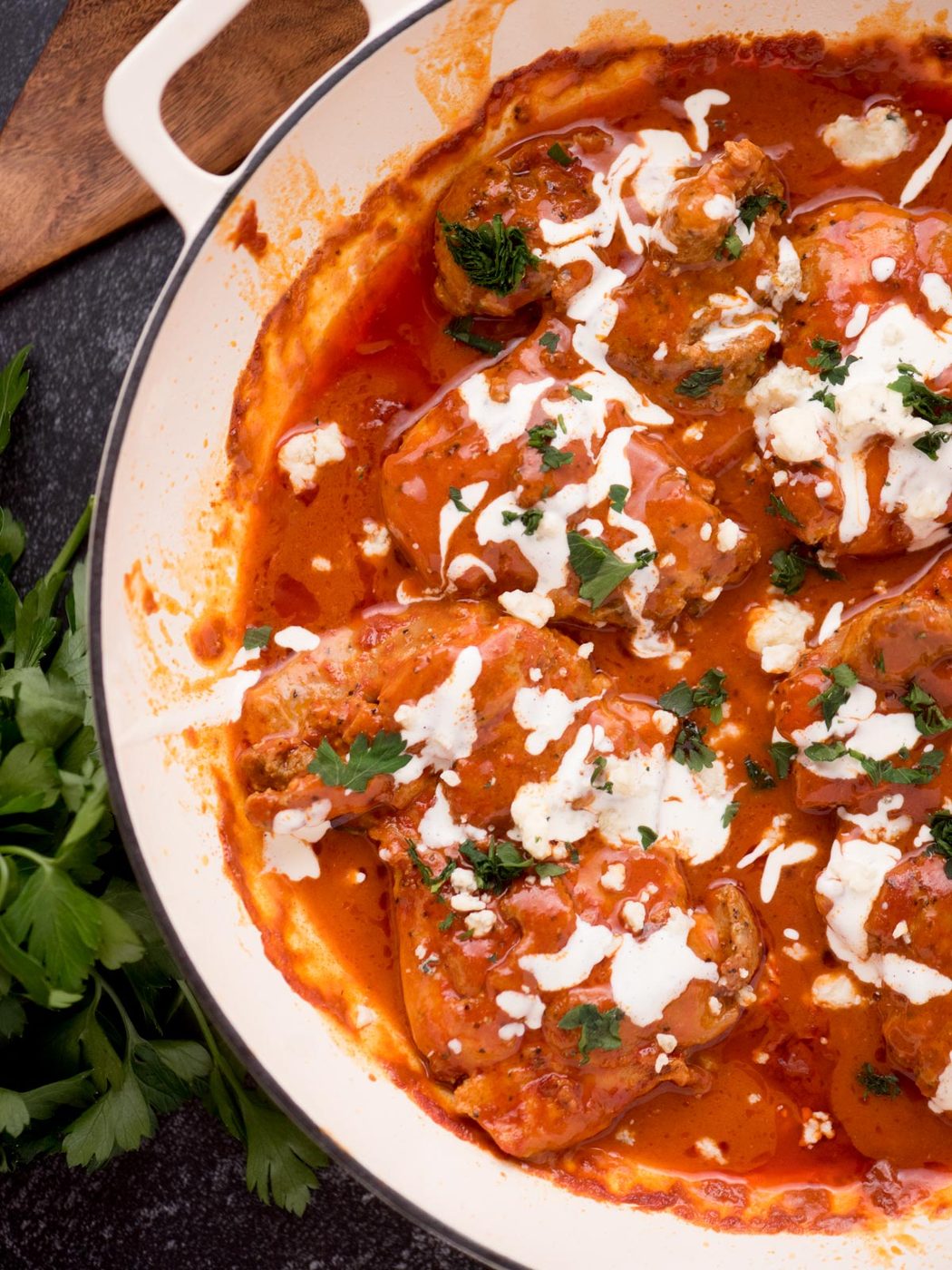 buffalo chicken thighs in a skillet surrounded by fresh parsley, a serving utensil, and a kitchen towel
